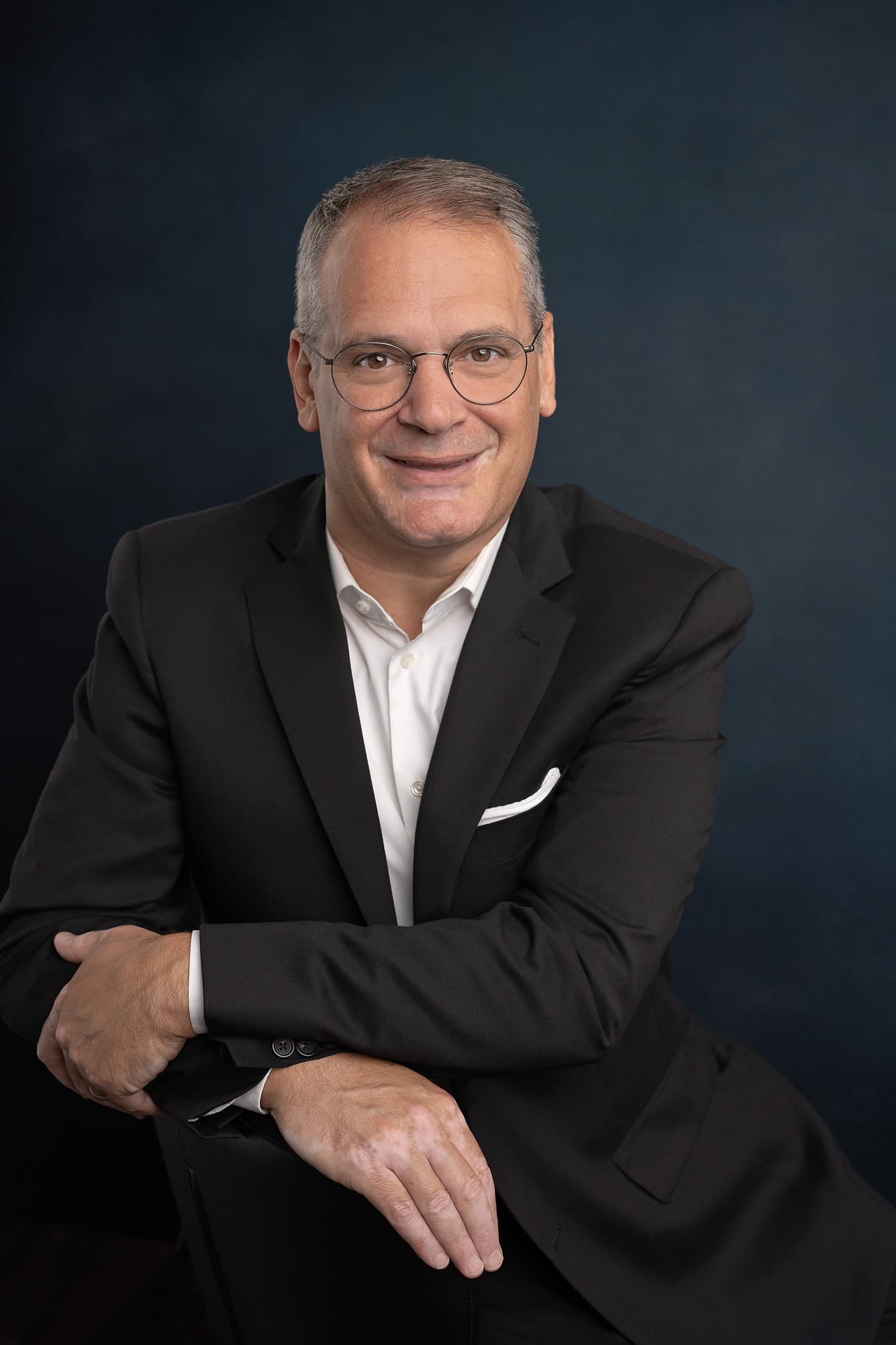 Stephen Golinski in a dark suit and white shirt, arms resting on a chair back, against a navy backdrop