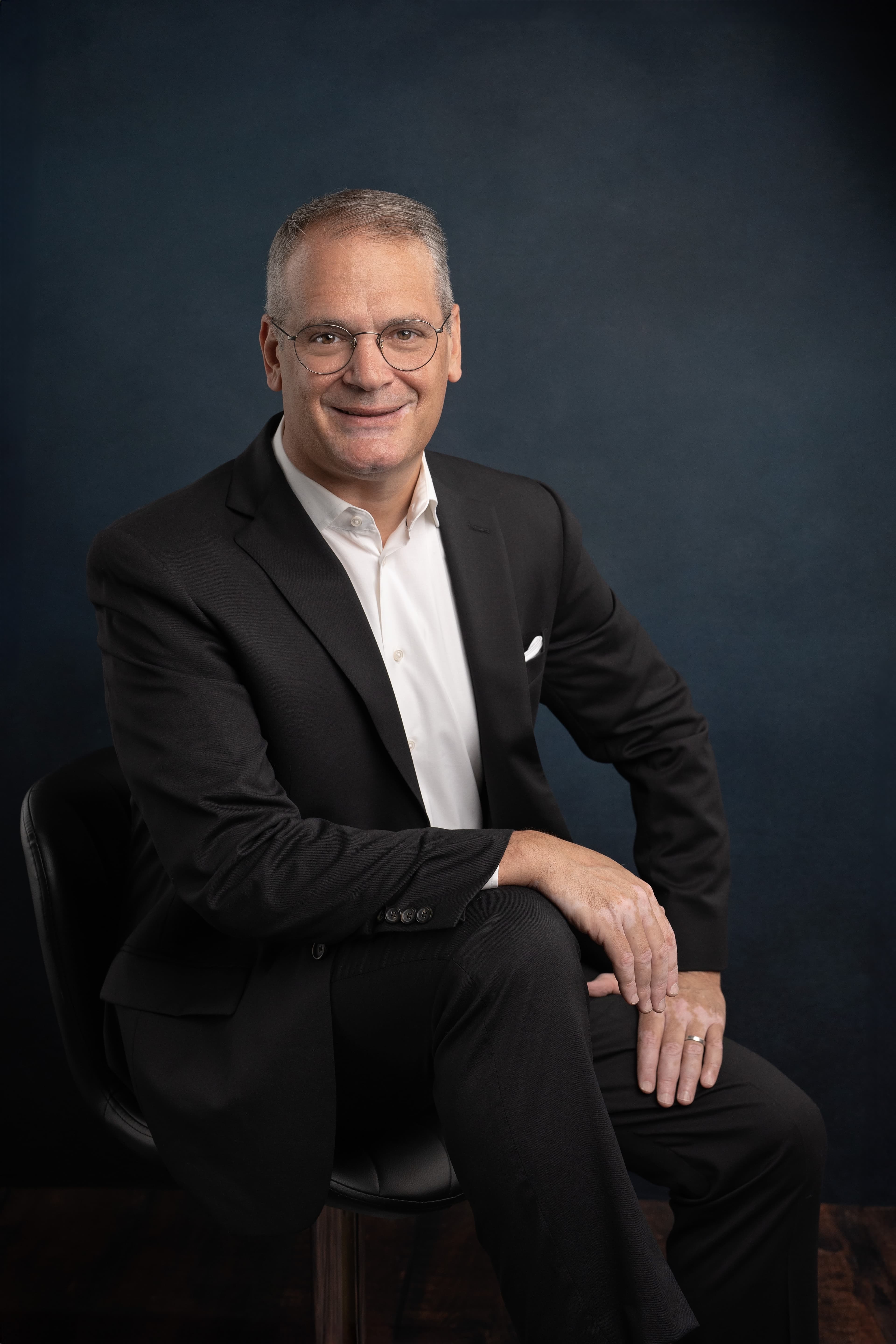Stephen Golinski seated in a dark suit against a navy backdrop, full length